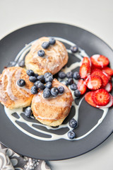 Cottage cheese pancakes or syrniki with blueberry and strawberries on black plate, closeup view. Condensed milk sauce. Russian, Ukrainian cuisine. Healthy tasty breakfast