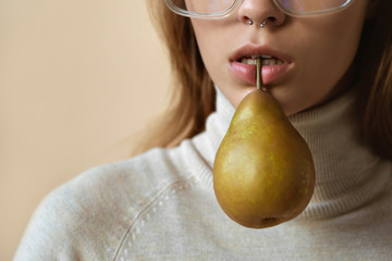 Fashion sexy young lady hold pear in mouth, stylish girl with piercing in nose wear trendy glasses, vogue attractive model in eyewear with fruit on beige studio background, copy space, close up view
