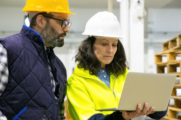 Two factory workers looking at laptop at plant. Confident technicians standing with modern device. Manufacturing concept