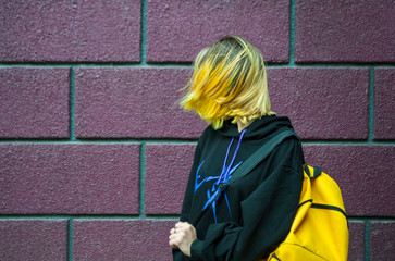 Cute teenager girl with yellow hair and backpack stand behind the brick wall