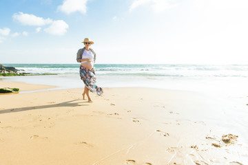 Happy pregnant woman holding hand on belly. Smiling future mother walking on beach during vacation. Pregnancy concept