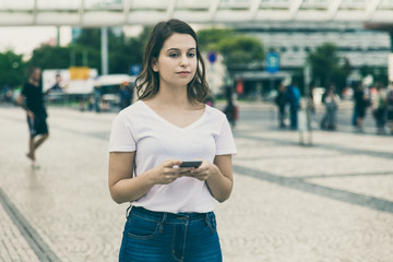 Fototapeta premium Front view of calm woman holding smartphone. Beautiful young lady using modern phone while standing on street. Technology concept