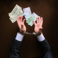 At the table sits a businessman in a black suit with handcuffs on his hands. Top view of hands with police handcuffs and currency on the table. Corruption, bribery, illegal transactions.