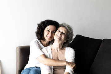 Happy cheerful middle aged woman hugging senior lady at home. Adult daughter sitting on couch and embracing senior mother. Family relations concept