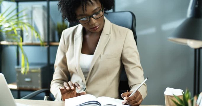 Close up portrait of thoughtful beautiful African American office worker writing down to notebook while sitting at desk indoor. Serious female professional making notes at workplace. Business concept