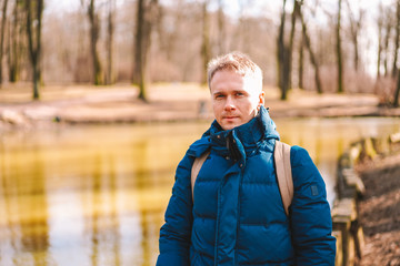 Fototapeta premium Portrait of a man in a warm jacket with a backpack in early spring in a nature Park near the lake