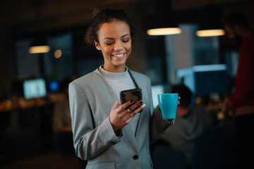 Young businesswoman using phone and drinking coffee in office.	