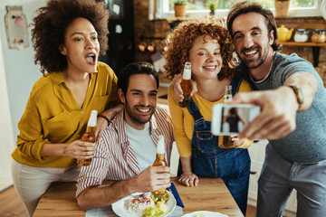 Happy friends drinking beer and taking selfie with smart phone in dining room.