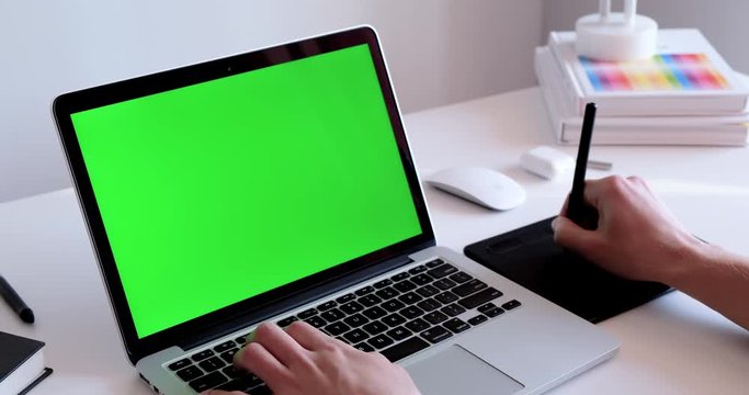 Close up of a man working on a laptop with a green screen chroma key display, drawing with a graphic tablet in a white office environment. Notebook mockup static video with hand typing on a keyboard 