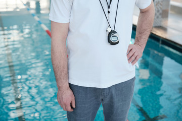 Close-up unrecognizable swimming coach with stopwatch and whistle on chest standing with hand on hip