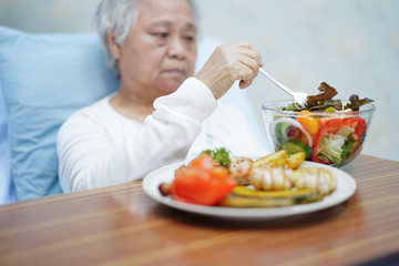 Asian senior or elderly old lady woman patient eating breakfast vegetable healthy food with hope and happy while sitting and hungry on bed in hospital.