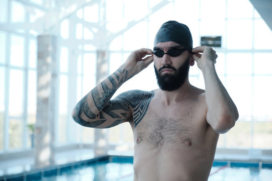 Bearded swimmer in black cap adjusting swimming goggles while standing at indoor pool