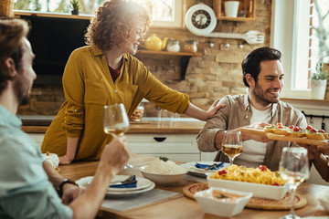 Happy friends talking and having fun while passing food at dining table.