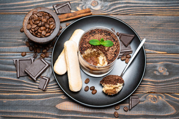 Classic tiramisu dessert in a glass cup, pieces of chocolate and savoiardi cookies on wooden background