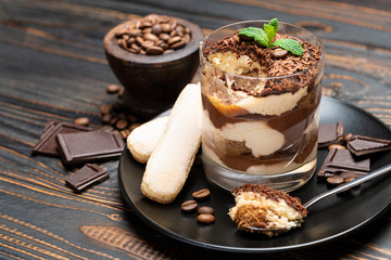 Classic tiramisu dessert in a glass cup, pieces of chocolate and savoiardi cookies on wooden background