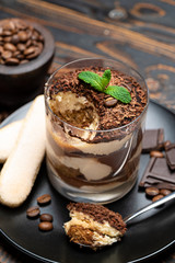 Classic tiramisu dessert in a glass cup, pieces of chocolate and savoiardi cookies on wooden background