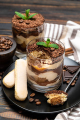 Classic tiramisu dessert in a glass cup, pieces of chocolate and savoiardi cookies on wooden background