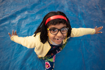 Low angle portrait of an Indian brunette baby girl with woolen clothes, cap and spectacles doing funny facial expression in a casual mood on a rooftop in winter afternoon. Indian lifestyle.