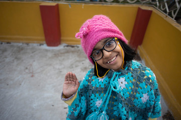 Low angle portrait of an Indian brunette baby girl with woolen clothes, cap and spectacles doing funny facial expression in a casual mood on a rooftop in winter afternoon. Indian lifestyle.