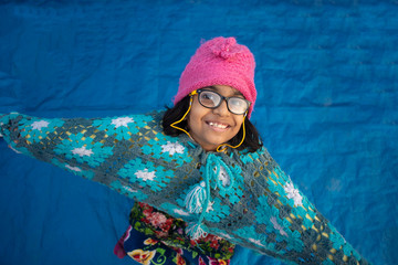 Low angle portrait of an Indian brunette baby girl with woolen clothes, cap and spectacles doing funny facial expression in a casual mood on a rooftop in winter afternoon. Indian lifestyle.