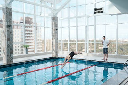 Swimming coach watching swimmer jumping in water, woman training with instructor in modern pool
