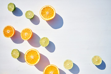 Ripe orange and lime halves loosely laid on white wooden background, shot from above in harsh sunlight with long colored shadows.