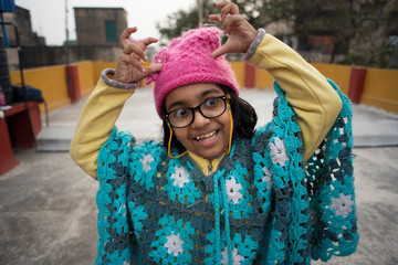 Low angle portrait of an Indian brunette baby girl with woolen clothes, cap and spectacles doing funny facial expression in a casual mood on a rooftop in winter afternoon. Indian lifestyle.
