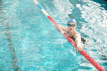 Portrait of healthy young woman in goggles and cap hanging on pool lane divider in swimming pool