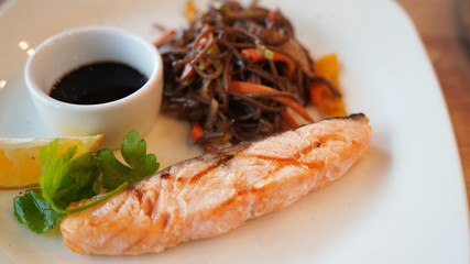 Close-up of delicious dish of buckwheat noodles with salmon and soy sauce with slice of lemon on white plate.