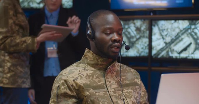 Close up of African American military worker in camouflage and headset talking via webcam while sitting in front of latop computer in monitoring room. Male operator at technical center speaking in mic