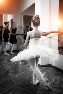 Young Ballet Dancer Posing By Ballet Bar, Stretching Right Leg Standing Back To The Camera Isolated In Studio Background. Small Ballerina Is Training At A Classical Ballet Studio. 