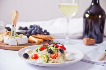 vegetables salad on the plate, bottle and glass of white wine and plate with assorted cheese, fruit and other snacks.