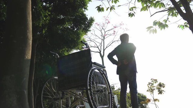 asian man old standing next to wheel chair stretching arms outdoors in park