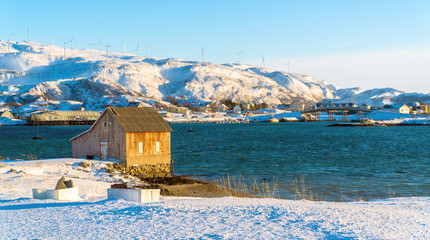 Norway. Old wooden house on the banks of a fjord in Sommarøy, with the Hillesøy Bridge in the background and windmills in the distance