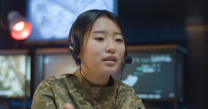 Asian young woman in uniform and headset sitting at computer screen in controlling room in army and talking via videochat. Close up of female soldier working in military data center.