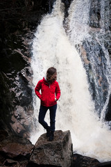 Frau steht in einer roten Jacke vor einem Wasserfall und schaut zur Seite