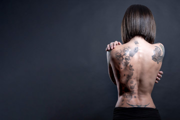 Donna con Schiena nudasi abbraccia con le mani con diversi tatuaggi , isolata su sfondo grigio scuro