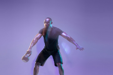 Fototapeta na wymiar Move. Young african-american bodybuilder training over purple background in neon, mixed light. Muscular male model with the weight. Concept of sport, bodybuilding, healthy lifestyle, motion and action