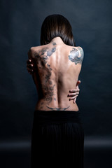 Fototapeta na wymiar Donna con Schiena nudasi abbraccia con le mani con diversi tatuaggi , isolata su sfondo grigio scuro