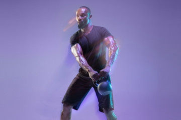 Move. Young african-american bodybuilder training over purple background in neon, mixed light. Muscular male model with the weight. Concept of sport, bodybuilding, healthy lifestyle, motion and action