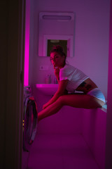 Sexy fashion young female wearing white lingerie posing in bathroom over neon pink and blue lights. Washing machine foreground. Technology concept. Party disco purple background