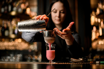 Fototapeta na wymiar brunette woman barman carefully pours cocktail from steel shaker into glass using sieve.