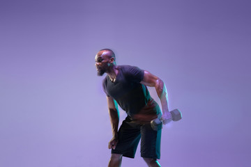 Emotions. Young african-american bodybuilder training over purple background in neon, mixed light. Muscular male model with weight. Concept of sport, bodybuilding, healthy lifestyle, motion and action