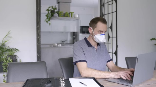 Man wearing sanitary surgical face mask and working on laptop while taking notes
