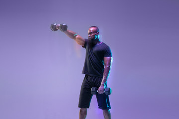 Emotions. Young african-american bodybuilder training over purple background in neon, mixed light. Muscular male model with weight. Concept of sport, bodybuilding, healthy lifestyle, motion and action