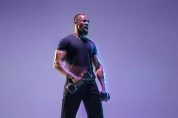 Emotional. Young african-american bodybuilder training over purple background in neon, mixed light. Muscular model with weight. Concept of sport, bodybuilding, healthy lifestyle, motion and action.