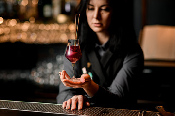 brunette woman bartender holds wineglass with red cocktail which decorated by two sticks.