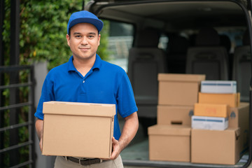 A smiling young asian delivery in blue uniform with parcel cardboard in front of customer house. Messenger and delivery concept.