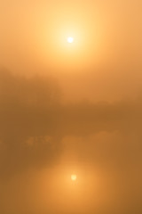 Beautiful peaceful sunrise or sunset countryside misty landscape of foggy water of river or lake, silhouettes of trees and soft suinlight transparenting through thick fog.