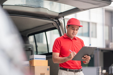 A smiling young asian delivery in red uniform with parcel cardboard in front of customer house. Messenger and delivery concept.